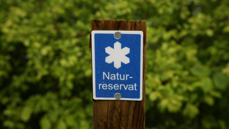 En blå och vit skylt för naturreservat sitter på en trästolpe med grönska i bakgrunden.