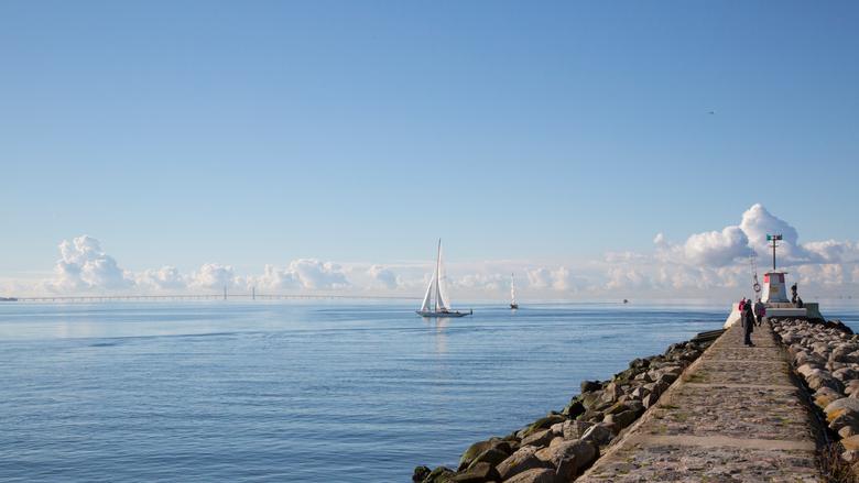 En segelbåt guppar på ett stilla hav, en pir av sten syns i ena kanten av bilden.
