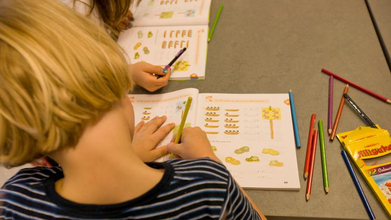 Ett barn med långt ljust hår syns bakifrån sittandes i en skolbänk och skrivandes i en mattebok.
