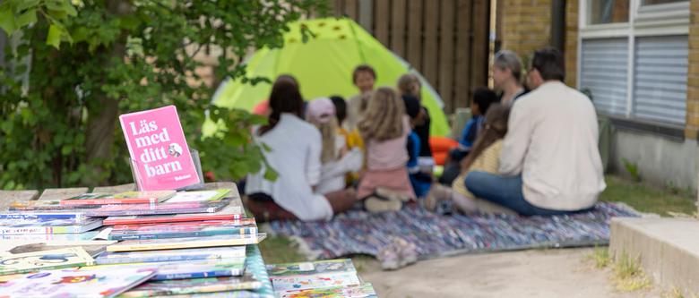 Böcker ligger på ett bord, en med titeln "Läs med ditt barn". I bakgrunden sitter barn och vårdnadshavare och lyssnar på en dramatiserad saga.