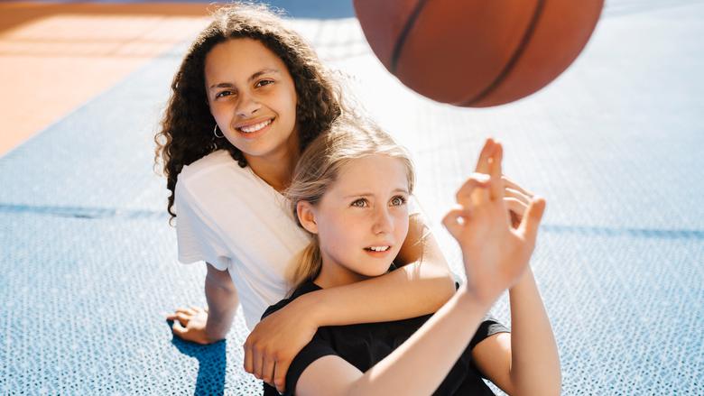 Två tjejer sitter utomhus på en basketplan och den ena snurrar en basketboll på fingrarna.