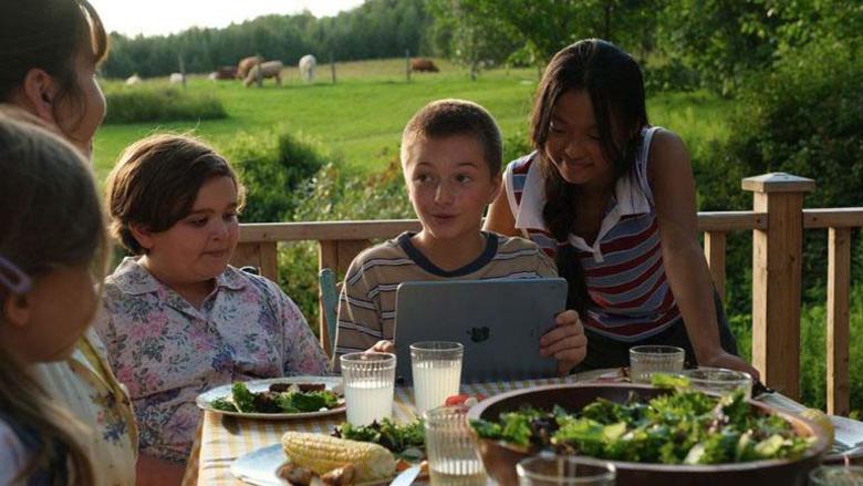 Några barn sitter samladerunt ett middagsbord utomhus på sommaren. Ett av barnen sitter med en laptop och några av de andra barnen tittar på vad som visas på skärmen.