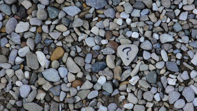 Bland hundratals stenar på en strand ligger en sten med ett frågetecken skrivet i svart på.
