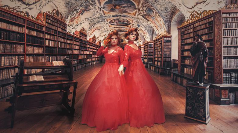 Två personer med röda långa klänningar och röda peruker poserar i ett gammalt bibliotek.