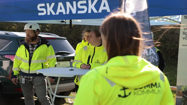 Personer i varselkläder står vid ett informationsbord under ett tält med texten Skanska. I förgrunden syns en person med jacka märkt Lomma kommun. Parkerade bilar och informationsmaterial finns i bakgrunden.
