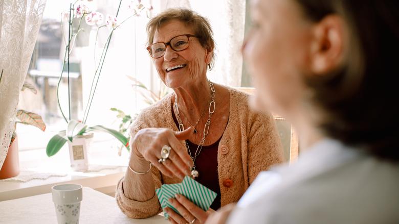 Äldre kvinna med kort rödlätt hår och kraftigt bågade glasögon sitter vid ett bord med spelkort i handen och ler.