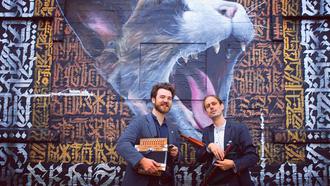 Två män i skjorta och kavaj står framför en väggmålning med motiv av en katt som öppnar sitt gap.