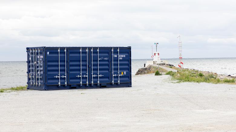En stängd container står uppställd på en grusyta med en pir, en liten fyr och havet i bakgrunden