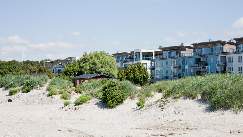 En rad lägenhetshus i kulörta färger syns bakom sanddyner med gröna vassdungar på toppen. 