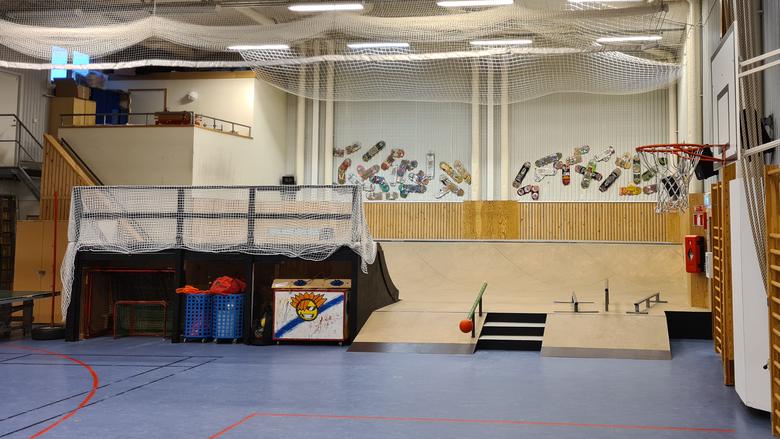 Översiktsbild över skejtrampen som är belägen i ena änden av en idrottshall. Skejtboardar pryder väggen bakom rampen som är ett par meter hög.