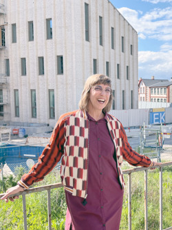 Person står vid ett räcke framför en byggarbetsplats med ett större tegelklätt hus under uppförande i bakgrunden. Personen bär en mönstrad jacka över en vinröd skjortklänning. Blå himmel med lätta moln syns ovanför byggnaden.