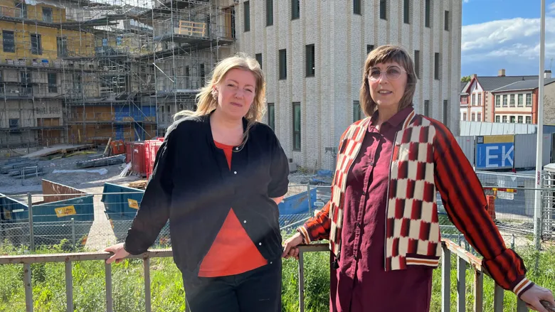 Två personer står vid ett räcke framför en byggarbetsplats. I bakgrunden syns en större byggnad under uppförande med ställningar, byggstaket och arbetsområde. Soligt väder med blå himmel och moln.