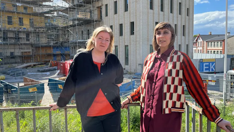 Två personer står vid ett räcke framför en byggarbetsplats. I bakgrunden syns en större byggnad under uppförande med ställningar, byggstaket och arbetsområde. Soligt väder med blå himmel och moln.