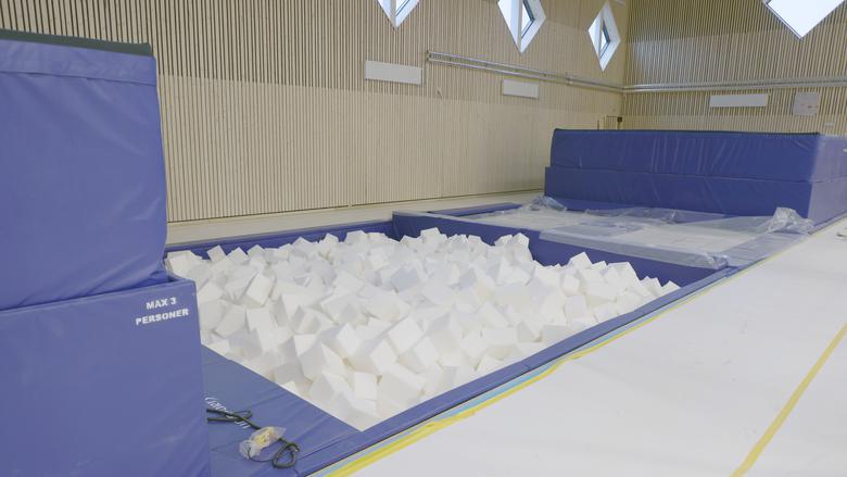 En massa vita mjuka kvadratiska skumgummibitar ligger i golvhöjd med höga blå madrasser intill. Bilden visar utrustning för gymnaster att träna hopp. 